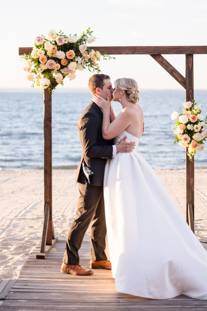 Bride and groom share their first kiss beneath a floral arch at their Crescent Beach Club beach wedding, with golden light and Long Island Sound as the backdrop for their September ceremony.