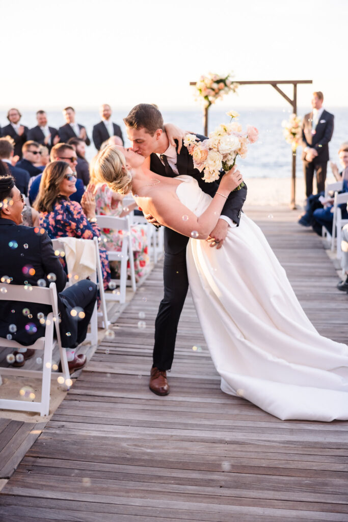 Groom dips and kisses his bride surrounded by guests and bubbles during their joyful recessional at a Crescent Beach Club September wedding on the Long Island waterfront.