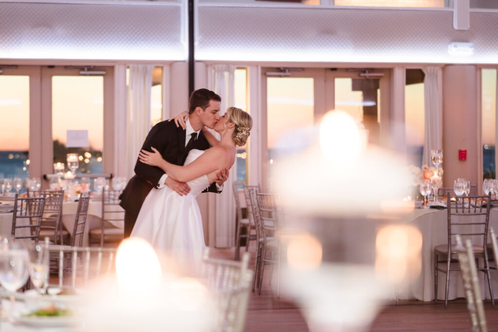 Bride and groom share a romantic kiss during their first dance in the candlelit reception space at Crescent Beach Club, framed by glowing lights and a Long Island sunset.