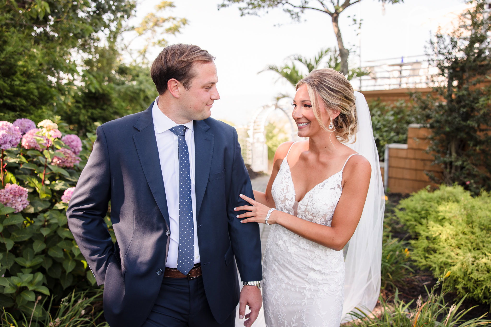 Bride and groom walk together during their first look along a garden path at a Crescent Beach Club waterfront wedding, sharing a relaxed moment surrounded by lush greenery.