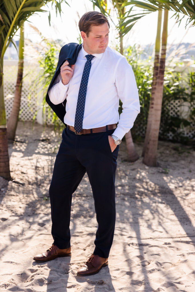 Groom stands beneath palm trees on the beach at a Crescent Beach Club waterfront wedding, holding his jacket over his shoulder in natural light before the ceremony.
