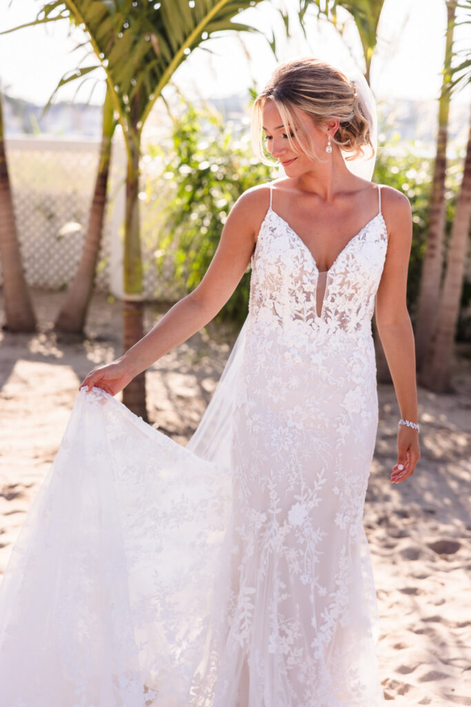 Bride in a lace wedding dress adjusts her gown beneath palm trees during portraits at a Crescent Beach Club waterfront wedding, with soft afternoon light and beach surroundings.