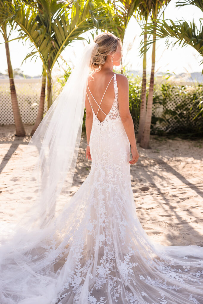 Back view of the bride’s lace wedding dress and flowing veil during beach portraits beneath palm trees at a Crescent Beach Club waterfront wedding.