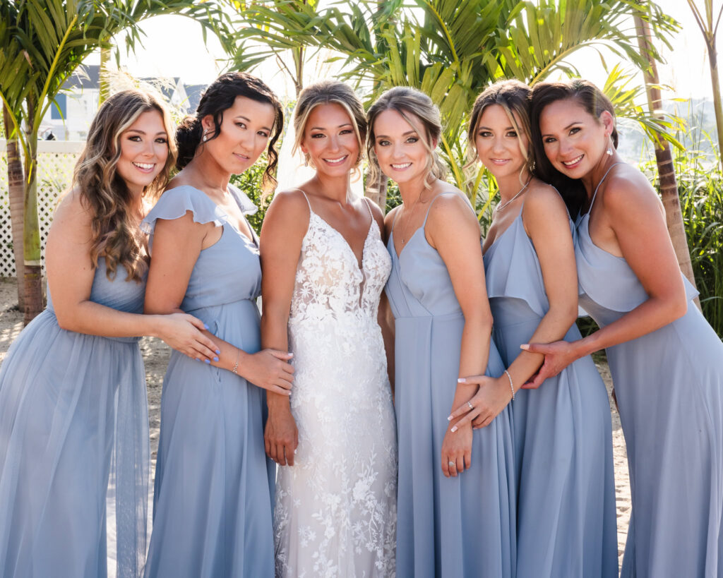 Bride poses with her bridesmaids in soft blue dresses during beach portraits at a Crescent Beach Club waterfront wedding, framed by palm trees and warm afternoon light.
