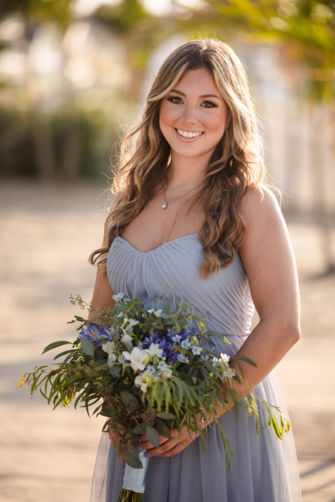 Bridesmaid in a soft blue dress holds a bouquet during golden light portraits at a Crescent Beach Club waterfront wedding, with palm trees softly blurred in the background.