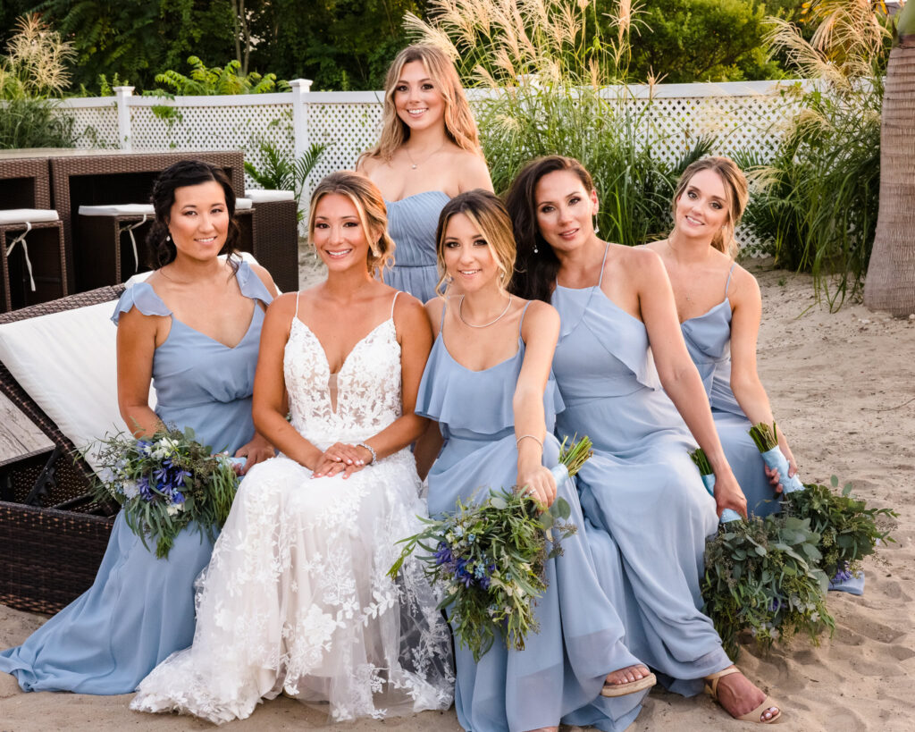 Bride sits with her bridesmaids in soft blue dresses during beach portraits at a Crescent Beach Club waterfront wedding, holding bouquets with natural greenery and blue florals.