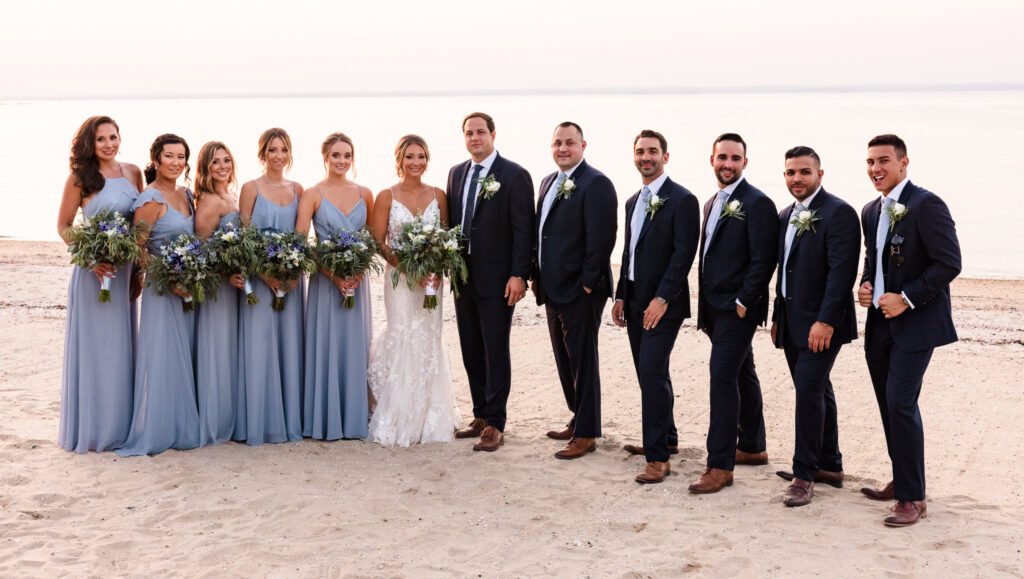Full wedding party stands together on the beach at a Crescent Beach Club waterfront wedding, with bridesmaids in blue dresses and groomsmen in navy suits against calm ocean views.