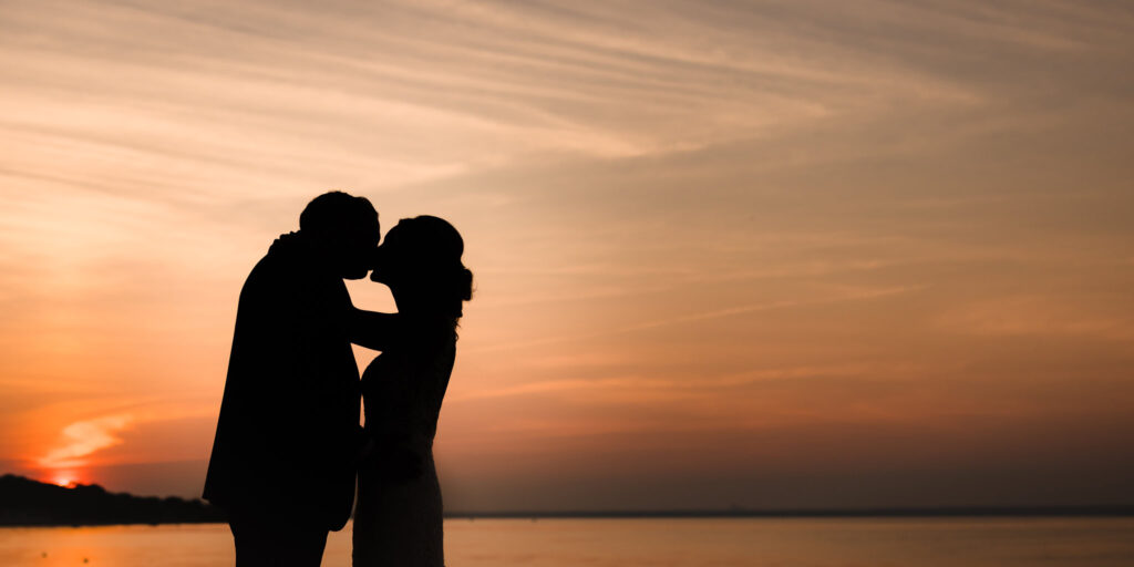 Silhouetted bride and groom kiss at sunset during a Crescent Beach Club waterfront wedding, with the sky glowing in warm orange tones over the water.