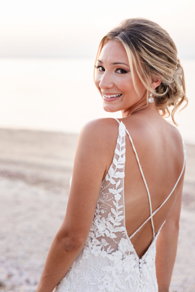 Bride smiles over her shoulder during beach portraits at a Crescent Beach Club waterfront wedding, highlighting the open-back lace detail of her gown against soft ocean light.