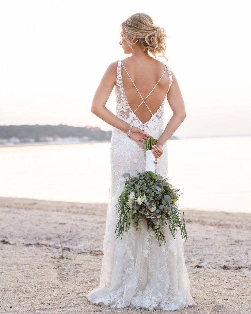 Bride holds her bouquet behind her back during sunset beach portraits at a Crescent Beach Club waterfront wedding, showcasing the open-back lace design of her gown. Send the next image when ready.
