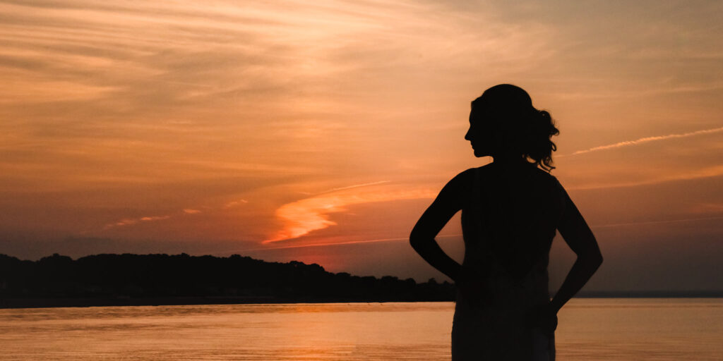 Silhouetted bride stands along the shoreline at sunset during a Crescent Beach Club waterfront wedding, with warm evening light reflecting across the water.