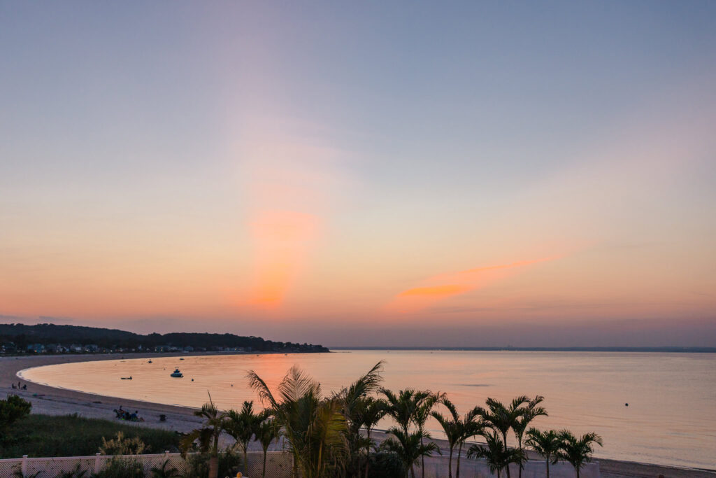 Sunset over Long Island Sound at a Crescent Beach Club waterfront wedding, with calm water, soft pastel skies, and palm trees lining the shoreline.