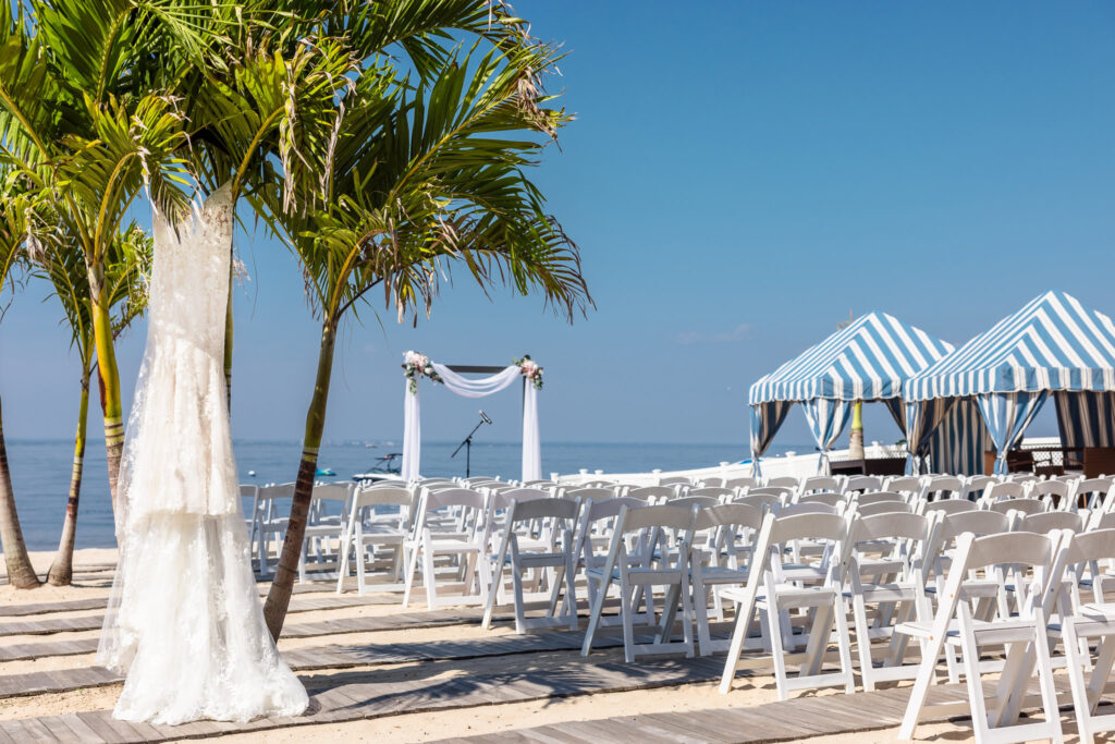 Lace wedding gown hanging between palm trees at Crescent Beach Club, overlooking a beachfront ceremony setup with white chairs and striped cabanas on Long Island Sound.