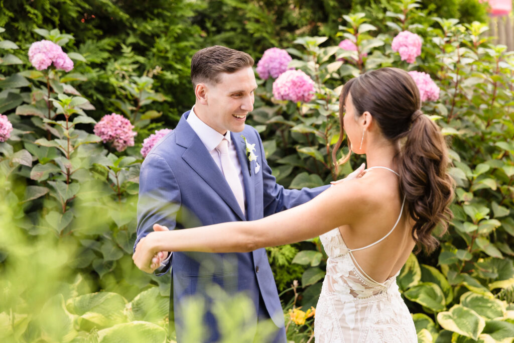 Emotional first look between bride and groom in a lush garden at Crescent Beach Club, surrounded by blooming pink hydrangeas and summer greenery