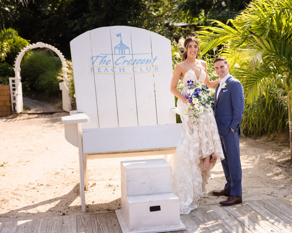 Bride and groom pose by the oversized white chair at Crescent Beach Club, with the bride holding a vibrant blue and white bouquet under tropical palm trees.
