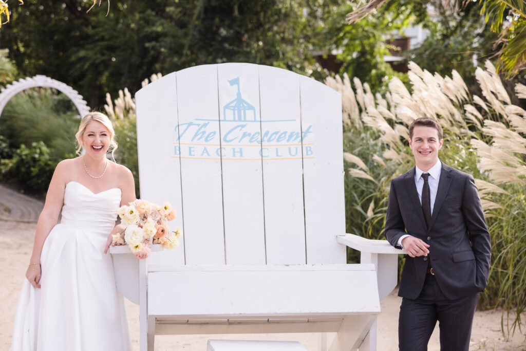 Joyful bride and groom pose beside the iconic Crescent Beach Club chair, surrounded by ornamental grasses and coastal greenery on the beachfront.