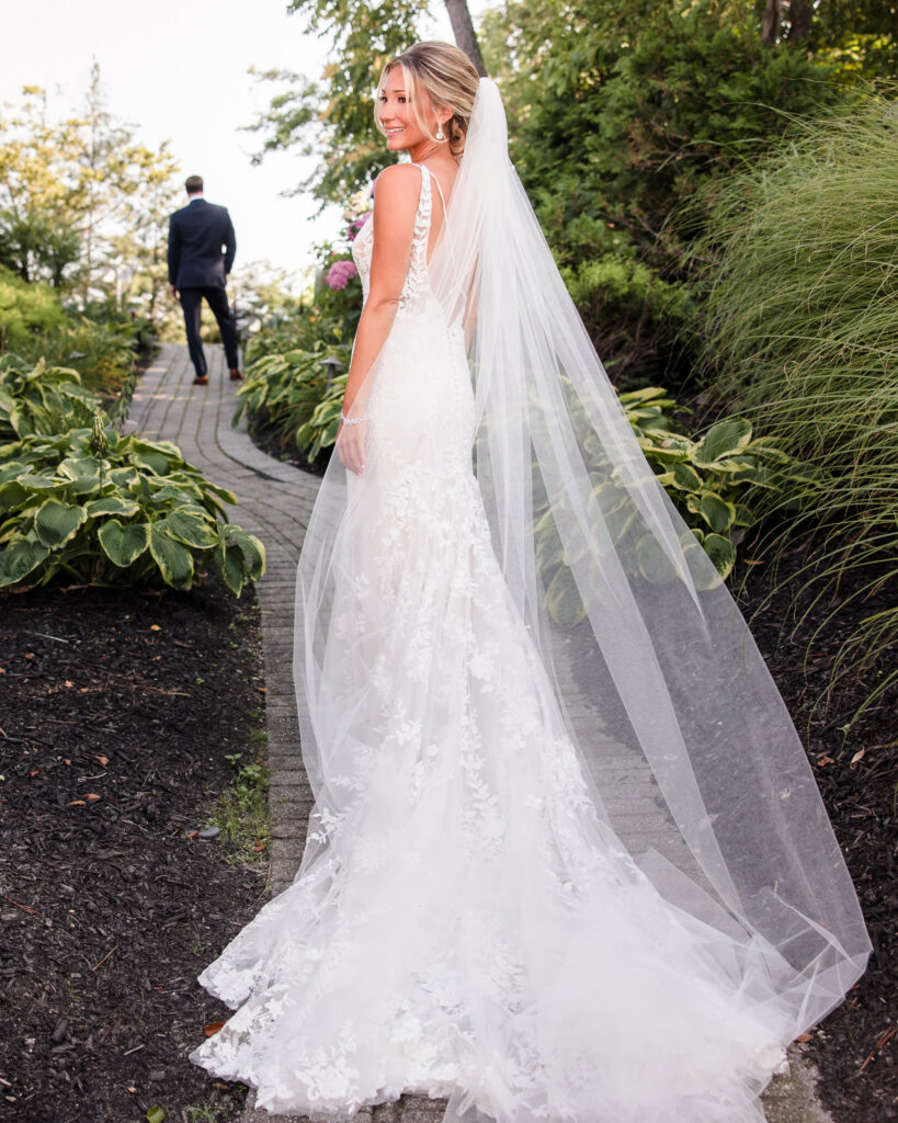 Bride in a lace wedding gown with a flowing veil smiles before a first look on the garden path at Crescent Beach Club, as the groom waits ahead.