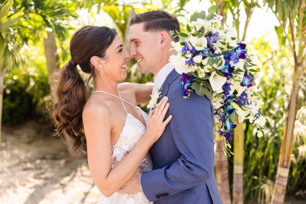 Bride and groom share a joyful, intimate moment under palm trees, with a cascading bouquet of white and vibrant blue flowers.