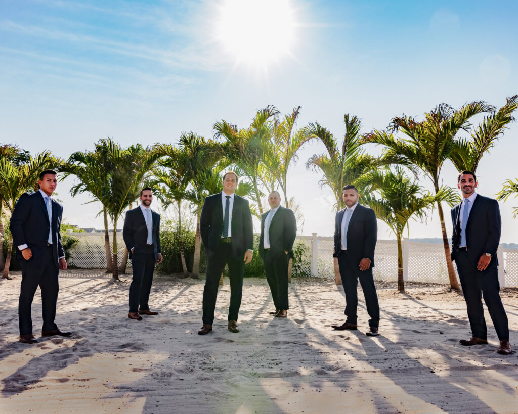 Groom and groomsmen pose confidently on the sand beneath palm trees, with the sun shining brightly overhead.