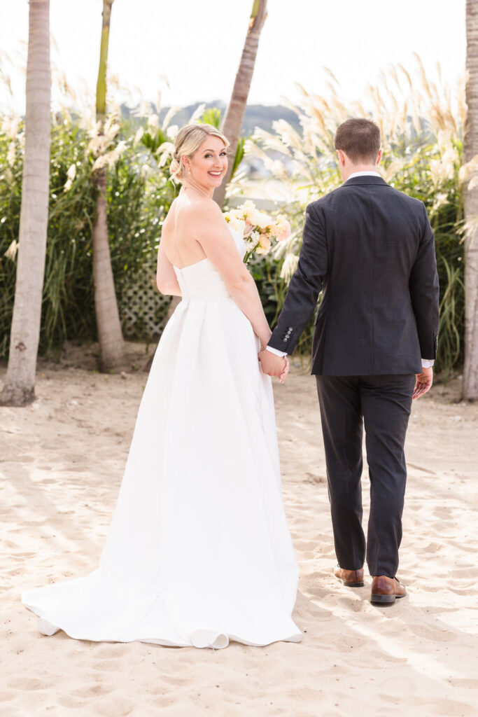 Bride smiling over her shoulder while walking hand-in-hand with her groom on the sandy beach at Crescent Beach Club, surrounded by palm trees and pampas grass.