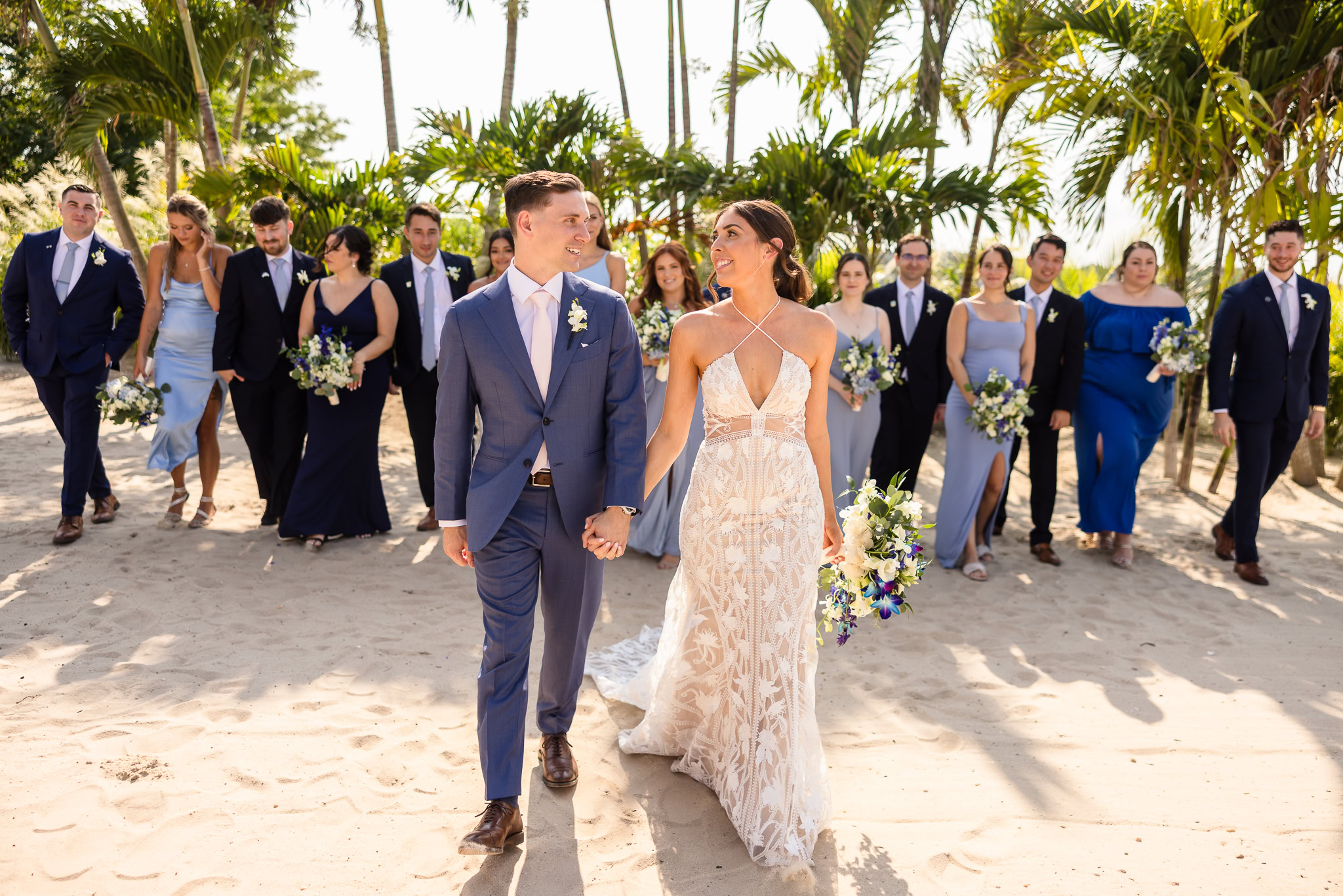 Bride and groom walk hand-in-hand across the sand at Crescent Beach Club, smiling at each other as their wedding party follows behind beneath swaying palm trees.