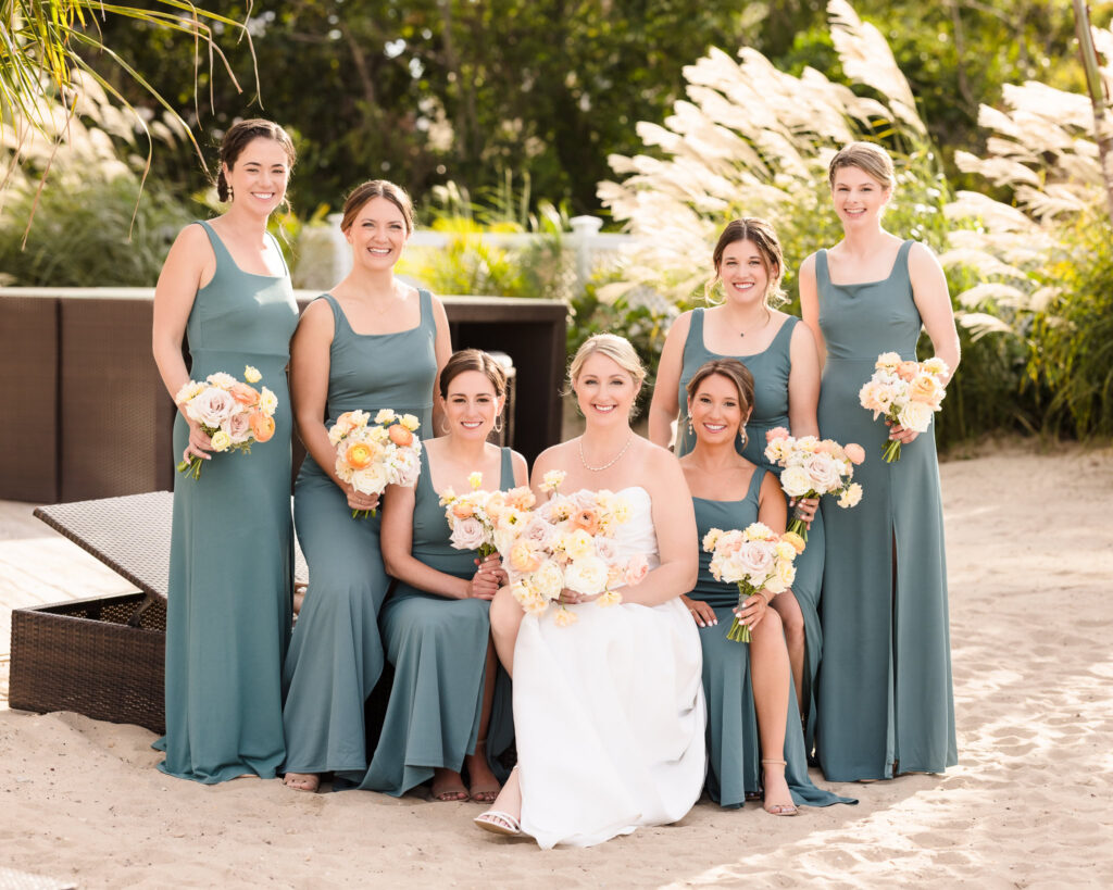 Bride poses with her bridesmaids on the sandy grounds, all holding pastel floral bouquets and smiling in the warm sunlight.
