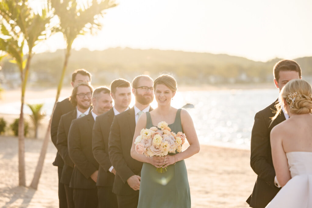 Bridesmaid holding a pastel bouquet smiles during a golden hour beachfront ceremony at Crescent Beach Club, with groomsmen lined up behind her in soft backlight.