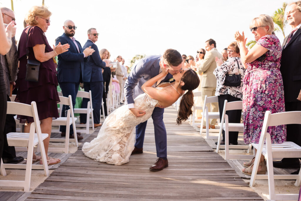 Groom dips and kisses the bride down the aisle as guests cheer and applaud during their joyful beach ceremony exit at Crescent Beach Club.
