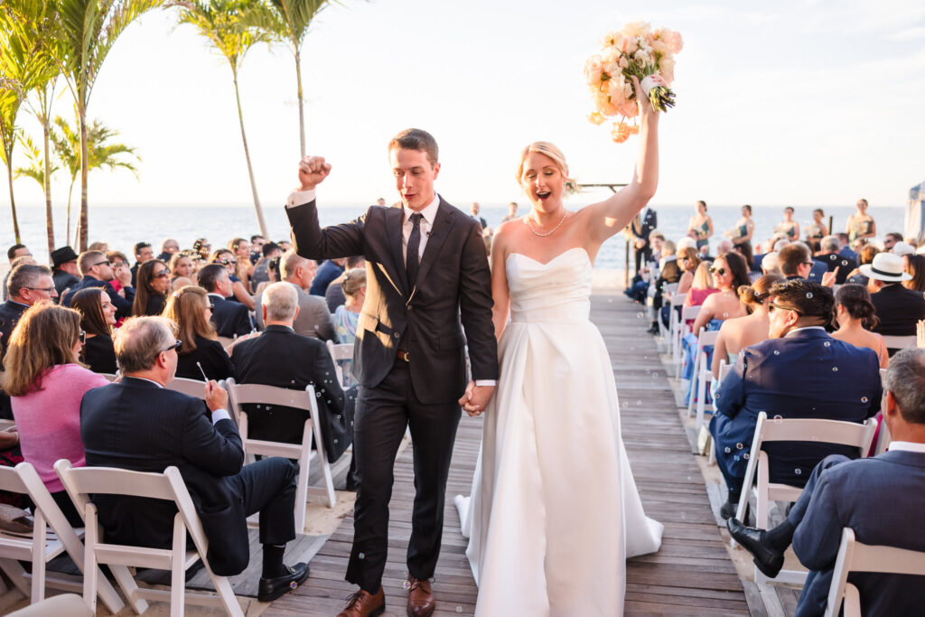 Joyful bride and groom celebrate their beachfront ceremony exit at Crescent Beach Club, surrounded by cheering guests and bubbles as they walk down the aisle.