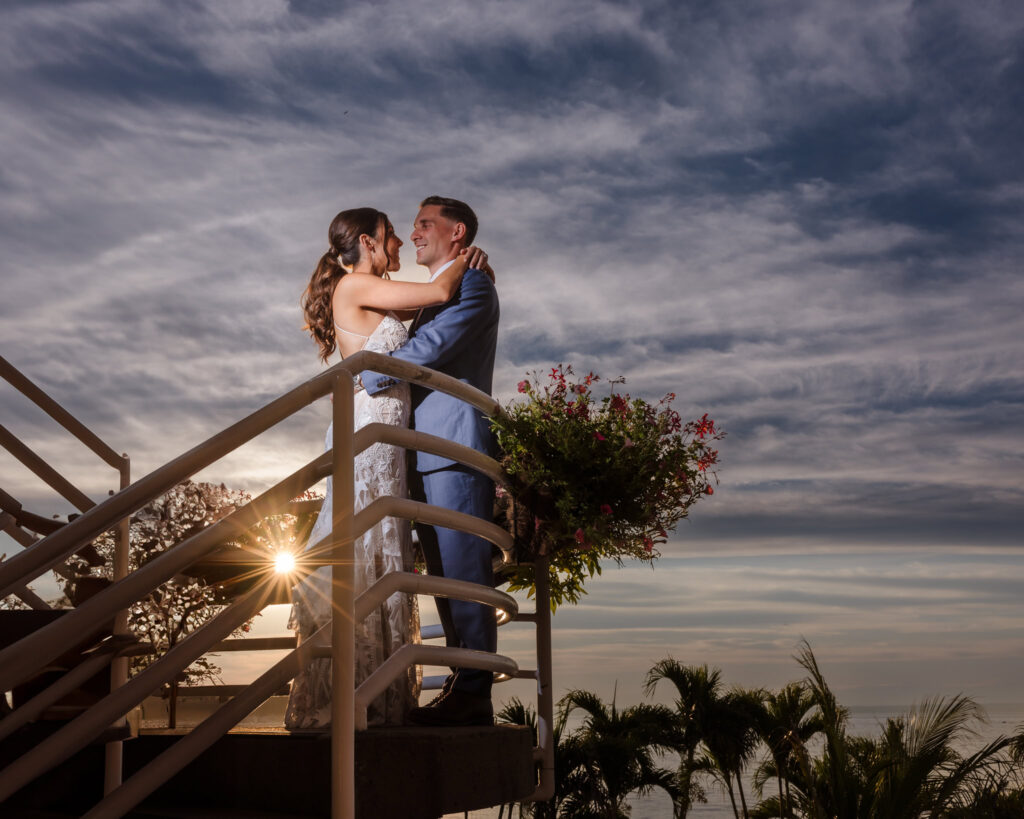 Bride and groom embrace on a staircase at Crescent Beach Club as the sun peeks through the railing, framed by dramatic skies, tropical flowers, and palm trees below.