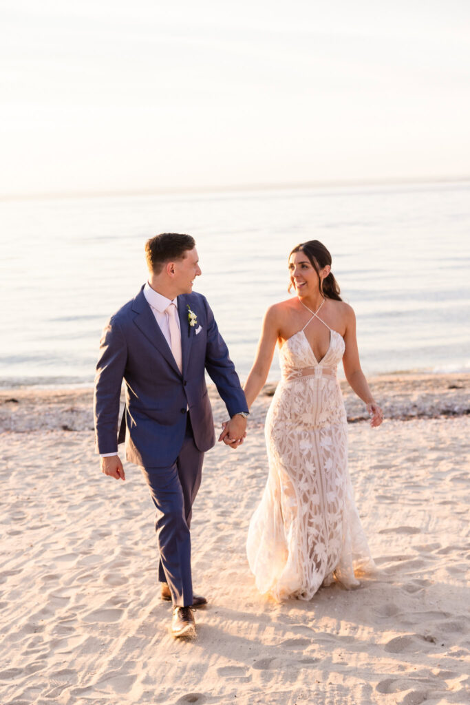 Bride and groom walk hand-in-hand along the shoreline, smiling at each other in the warm golden light of sunset.
