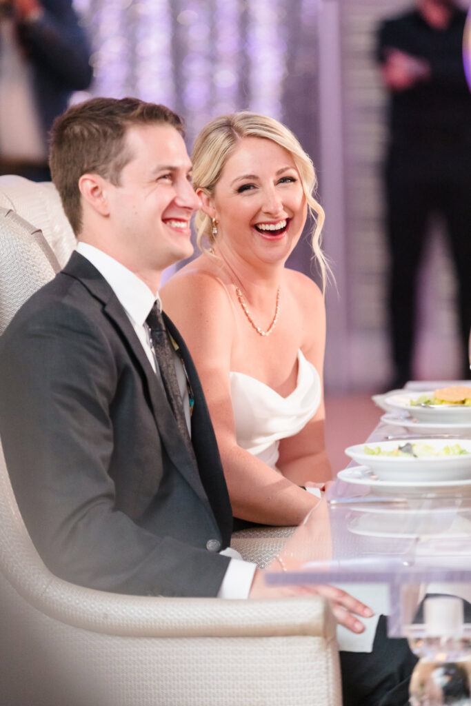 Bride and groom share a joyful laugh during their reception, seated together at their sweetheart table surrounded by candlelight and dinner plates.