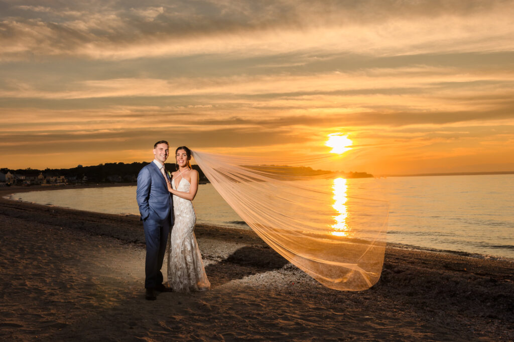Bride and groom pose on the beach at Crescent Beach Club with the bride’s veil flowing in the wind, framed by a breathtaking golden sunset over the Long Island Sound
