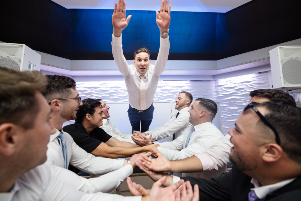 Groom dives into a tunnel of cheering friends' arms during a lively reception moment, surrounded by laughter and celebration.