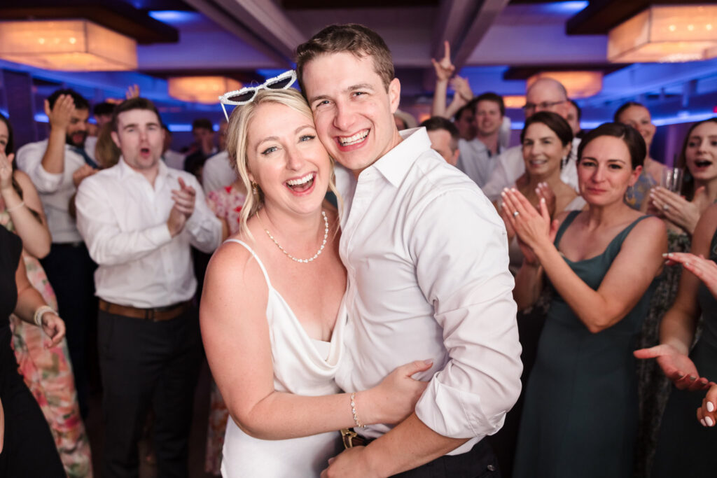 Bride and groom laugh and embrace on the dance floor at their reception, surrounded by cheering friends and lively celebration.