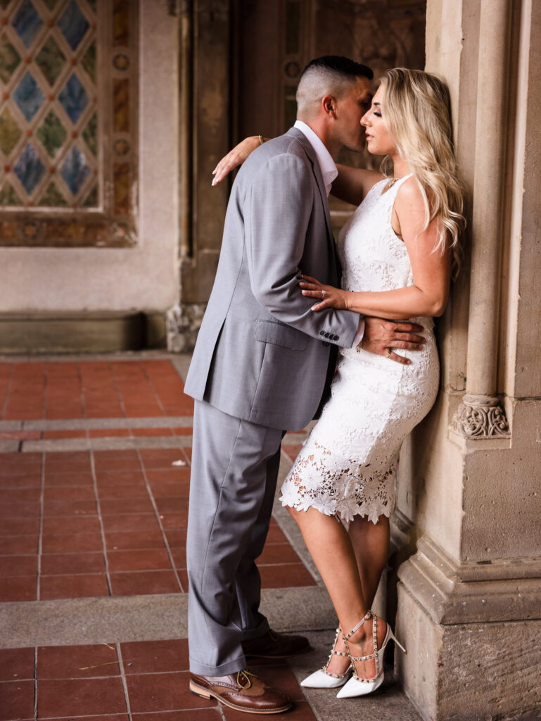Romantic editorial moment between engaged couple leaning against a column at Bethesda Terrace, styled in modern white lace and a grey suit for their Central Park engagement session.