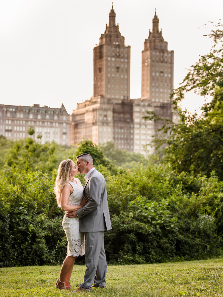 Couple shares a romantic kiss in Central Park with the iconic San Remo towers in the background during their fashion-inspired engagement session in NYC.