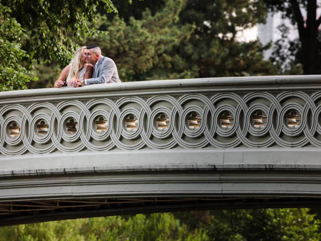 Engaged couple shares a kiss atop Bow Bridge, blending classic New York architecture with a romantic editorial engagement session in Central Park.