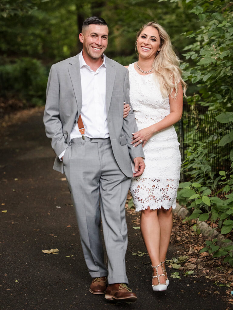 Couple walking arm in arm along a shaded Central Park path during their editorial engagement session, dressed in refined white and grey attire.