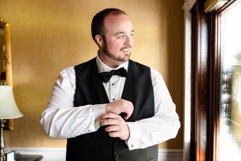 Groom smiling while adjusting his cufflinks near a window at a Long Island wedding venue, dressed in a classic black tuxedo with a bow tie.