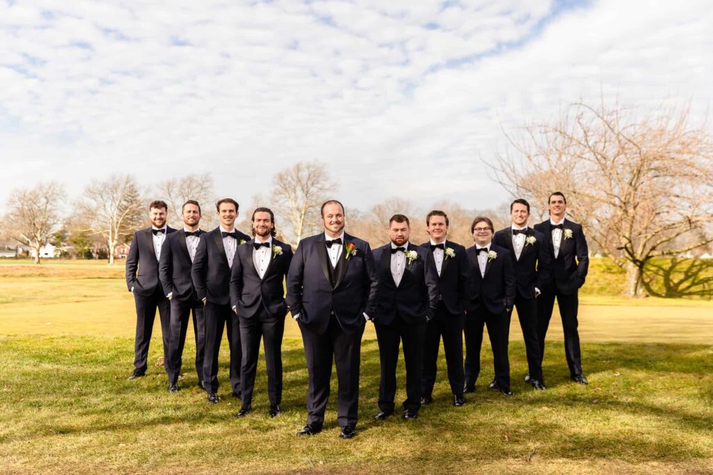 Groom standing proudly with his groomsmen in matching black tuxedos on a golf course, under a bright winter sky in Long Island at East Wind.