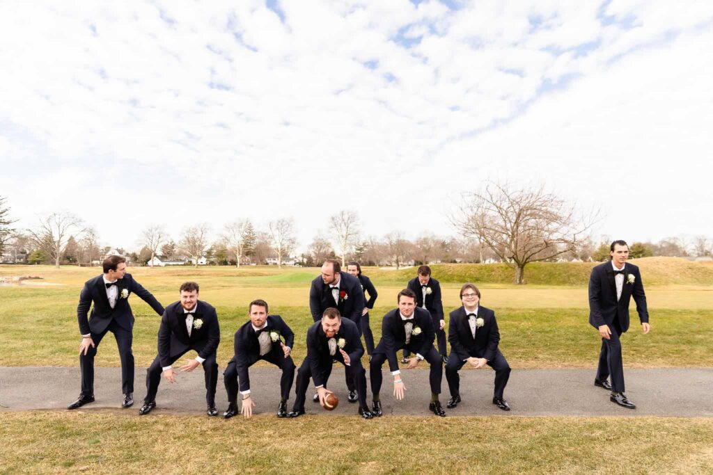 Groomsmen dressed in tuxedos posing in a playful football lineup with the groom as quarterback on a golf course during a Long Island wedding.