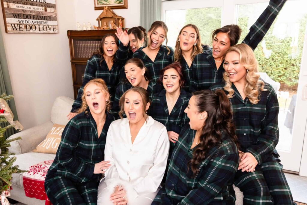 Bride in white pajamas laughing with her bridesmaids in matching plaid pajamas during joyful wedding morning preparations at a cozy Long Island home.