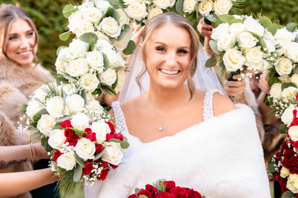 Radiant bride in a pearl-strapped gown and white fur wrap smiling as bridesmaids frame her with bouquets of ivory and red roses on her winter wedding day.