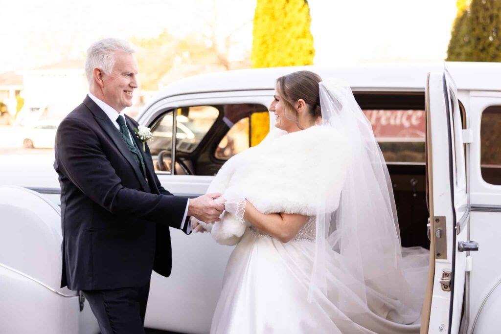 Bride stepping out of a vintage white car, smiling warmly as her father holds her hands before walking down the aisle on a winter wedding day at East Wind.