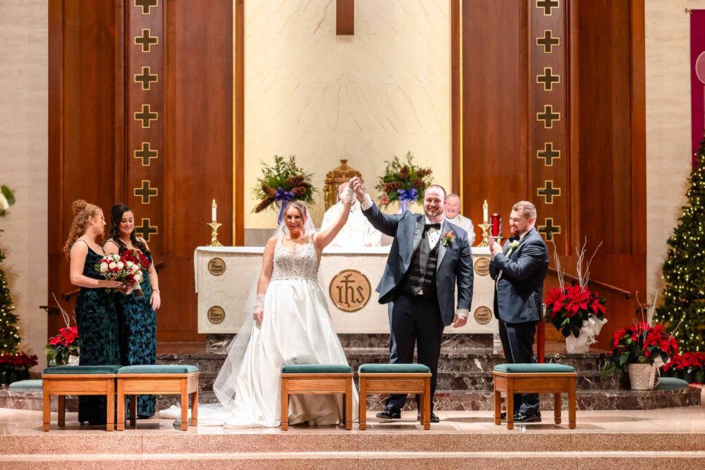 Bride and groom joyfully raising hands at the altar after being pronounced married in a Christmas-decorated Catholic church at East Wind, surrounded by their wedding party.
