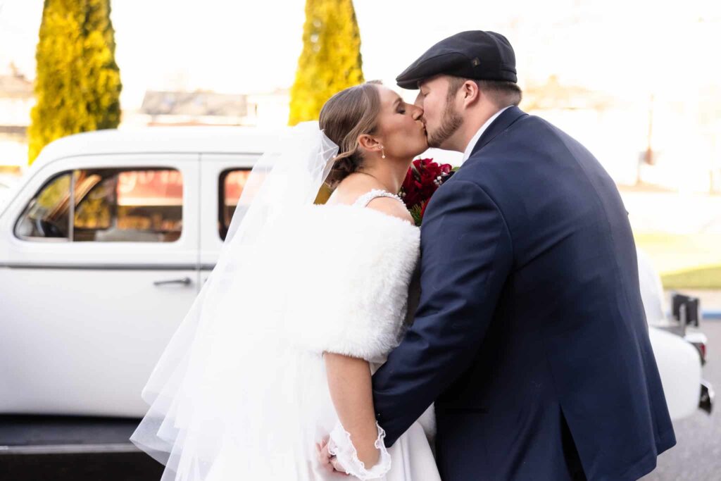 Bride and groom sharing a romantic kiss beside a vintage white car after their winter wedding ceremony at East Wind, with the groom in a navy suit and flat cap.