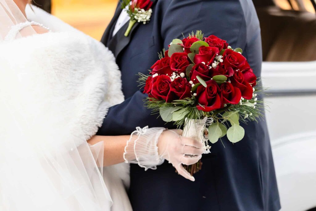 Close-up of bride’s hand in sheer gloves holding a romantic bouquet of red roses and winter greenery, embraced by the groom in a navy suit.