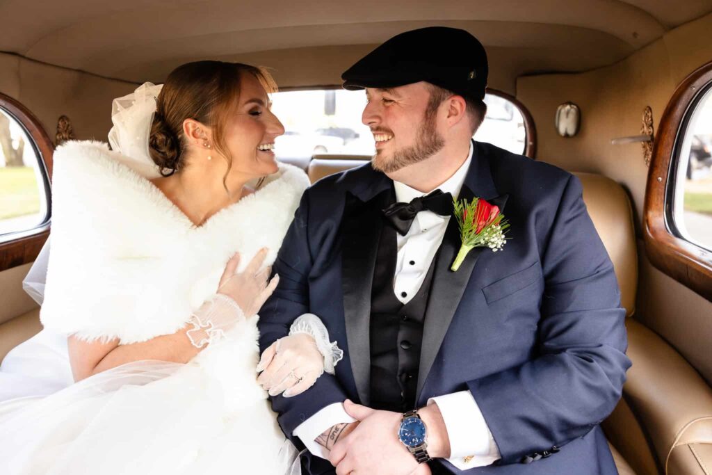 Bride and groom sharing a joyful moment in the backseat of a vintage car, dressed in winter wedding attire with festive boutonnière and faux fur wrap.