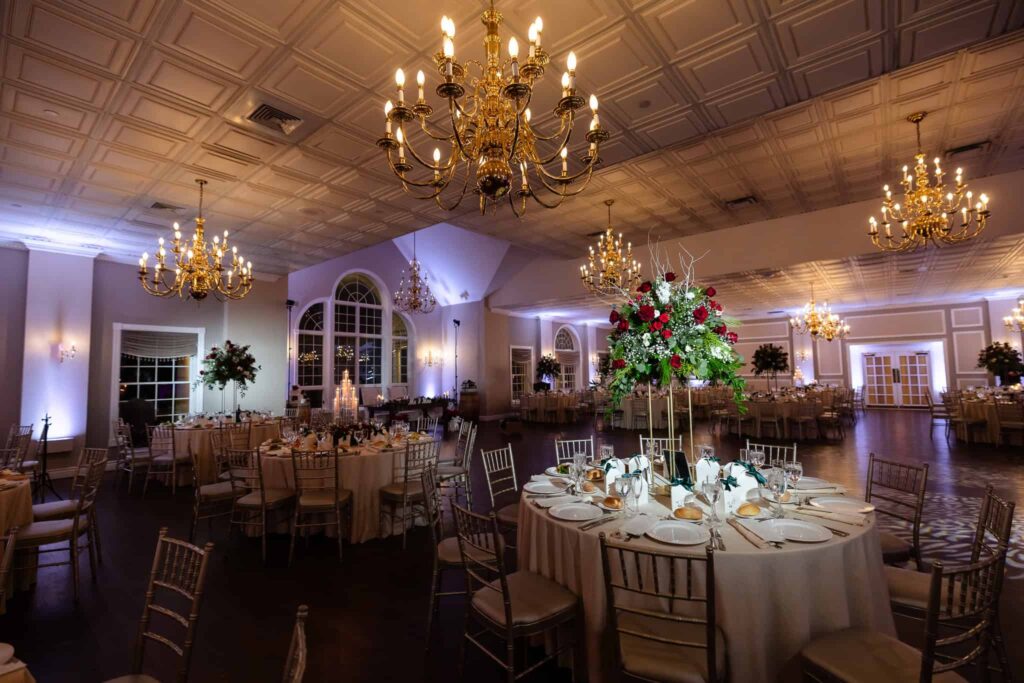 Elegant wedding reception ballroom with gold chandeliers, ivory linens, and tall floral centerpieces of red and white roses at a Long Island wedding venue at East Wind.